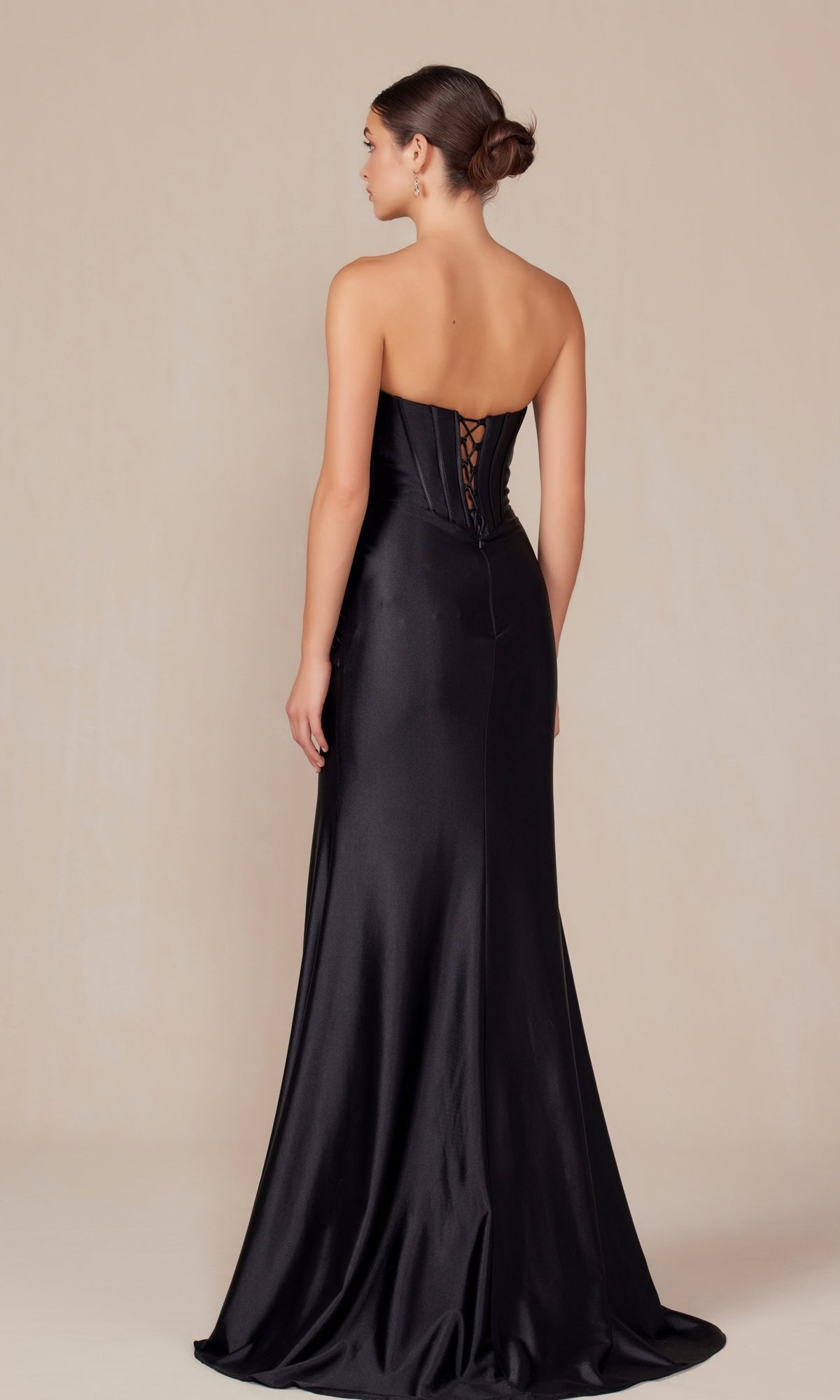 A woman stands facing away, wearing the Nox Anabel T1500—a strapless long corset prom dress featuring a floor-length black satin gown and lace-up detail on the bodice.